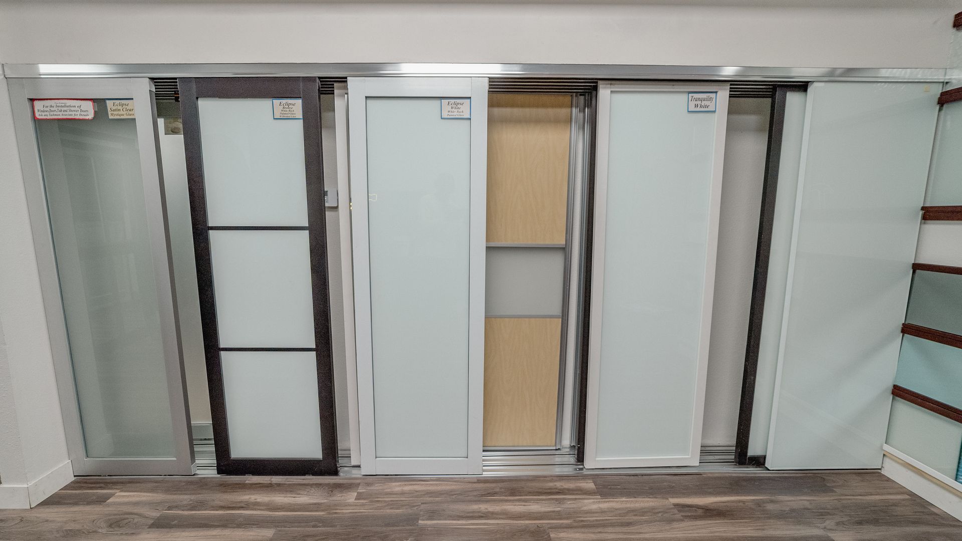 Sliding closet doors: dark brown, white, and frosted glass panels, displayed in a showroom with wooden floors.