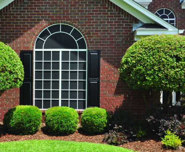 Brick house with large arched window and black shutters, surrounded by green bushes and landscaping.