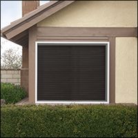 Dark window shutter on a beige house, with a green hedge in front.