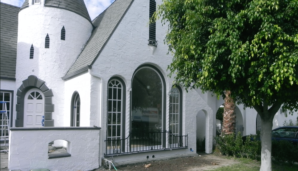 White stucco church with arched windows and entrance, partly shaded by a large tree