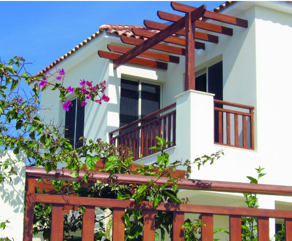 White house with wooden balcony and pergola, blue sky, surrounded by green and pink flowers.