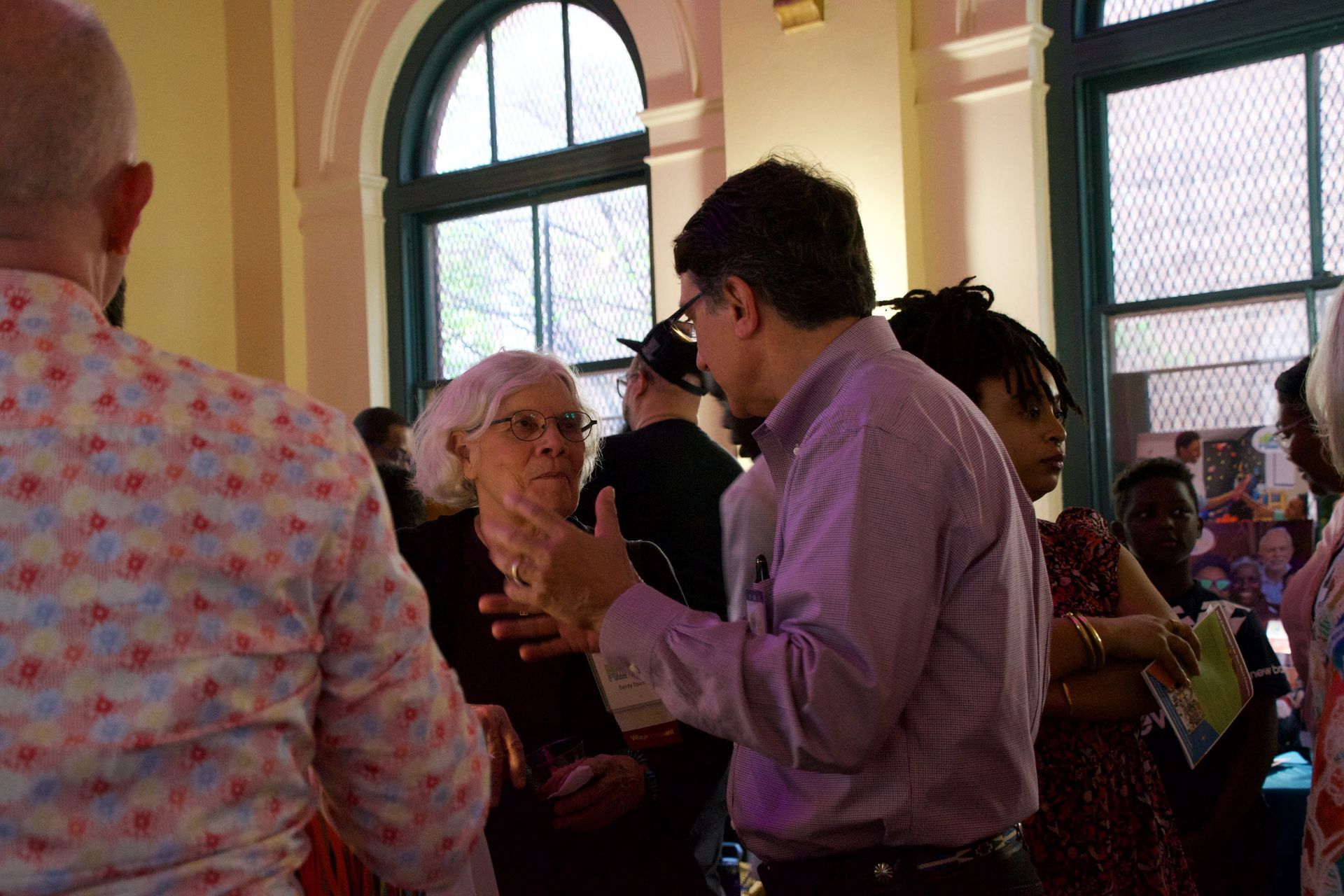 A group of people talking in a sunlit room with large arched windows, with one person gesturing while speaking.