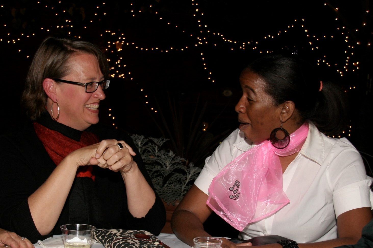 Two women are sitting at a table talking and one has a pink scarf around her neck