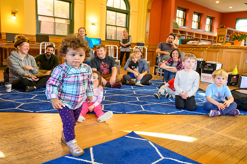 A little girl is walking in front of a group of people sitting on the floor.