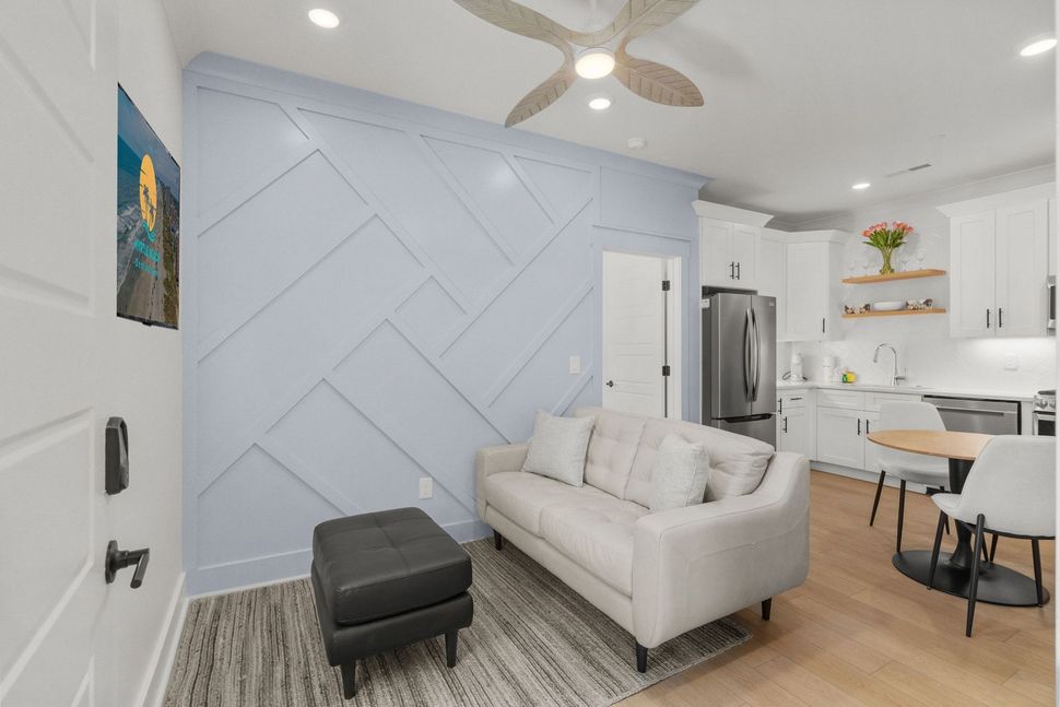 Bright living room with pale blue accent wall, cream sofa, black ottoman, and open kitchen with dining table