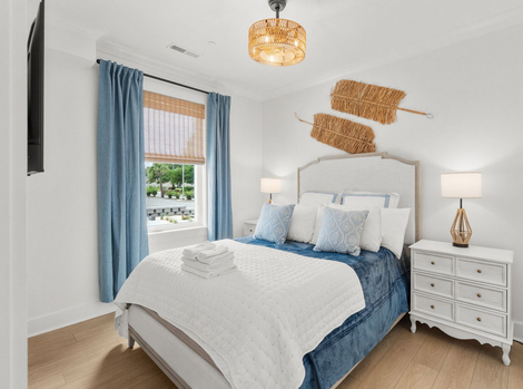Bright bedroom with white bed, blue bedding, and curtains beside a window with woven blinds