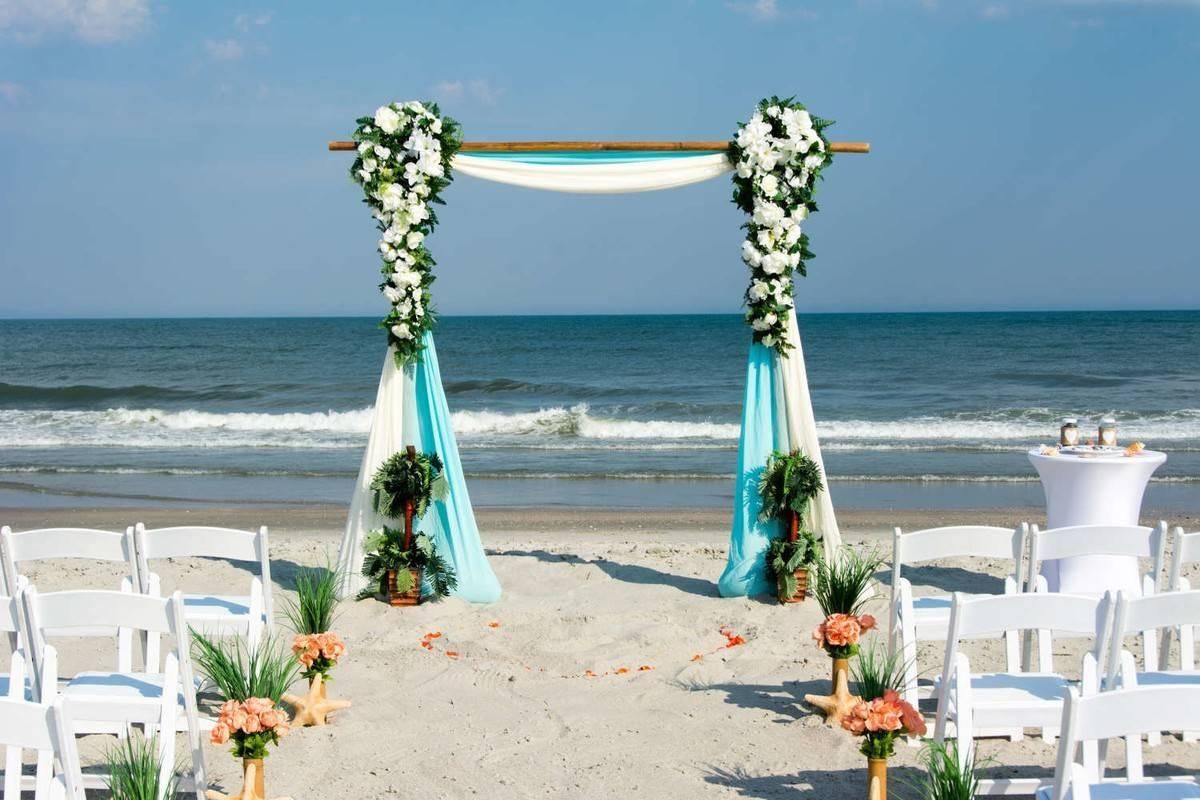 Beach wedding arch with white and turquoise floral decor facing the ocean, with rows of white chairs on sand