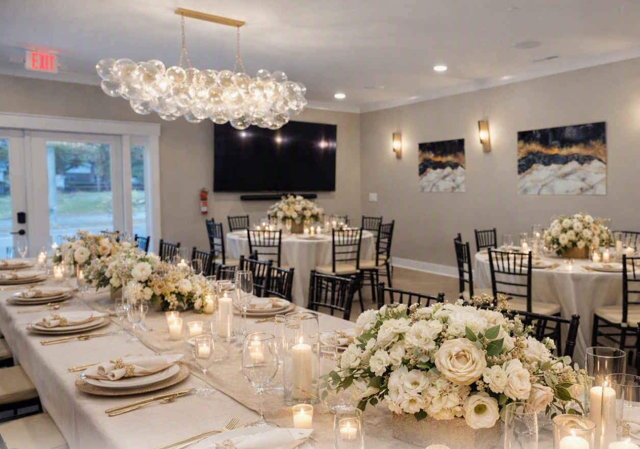 Elegant banquet hall with white floral centerpieces, set tables, chandeliers, and a wall-mounted TV