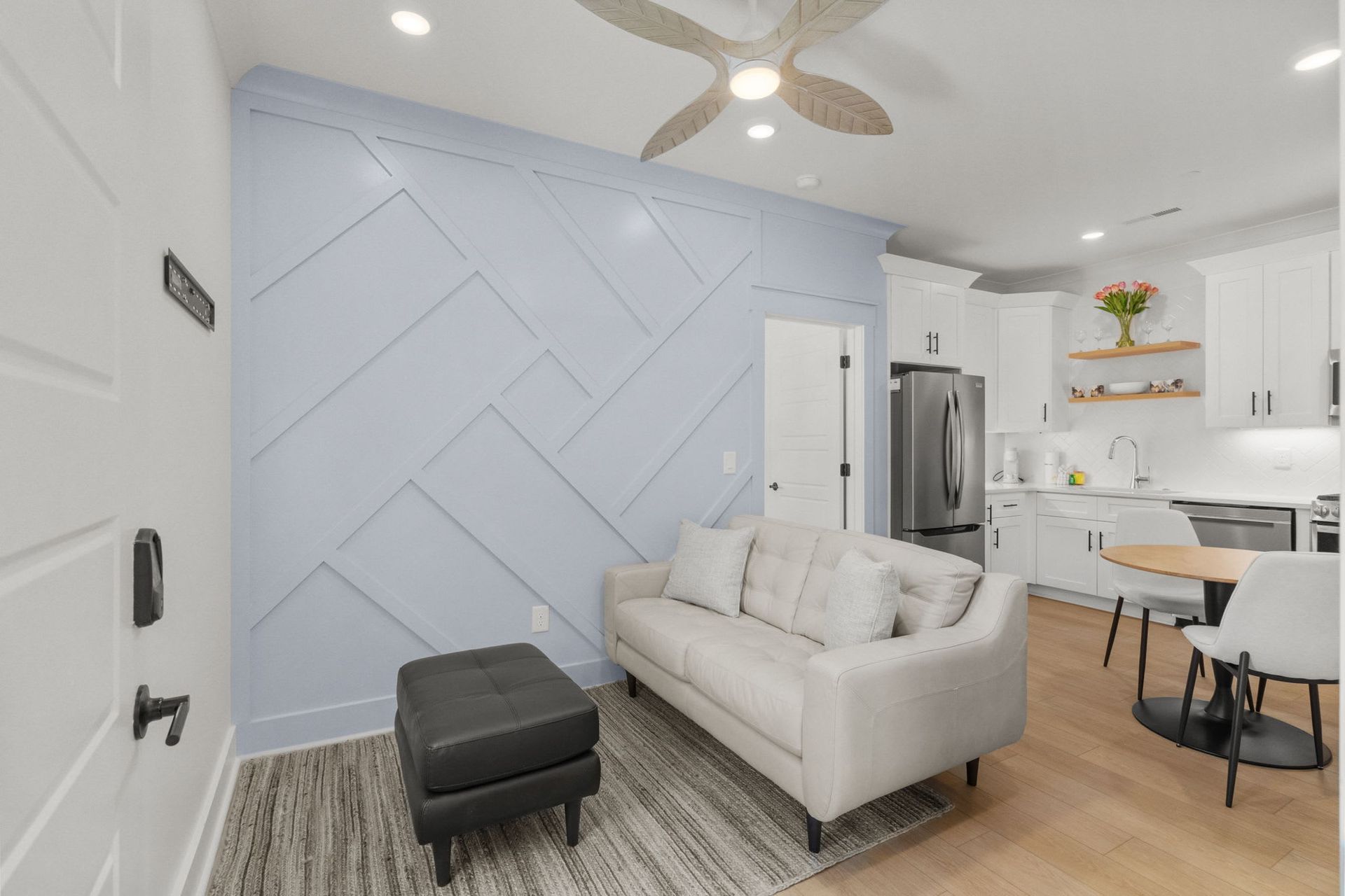 Bright living room with light blue accent wall, white sofa, black ottoman, and open kitchen area
