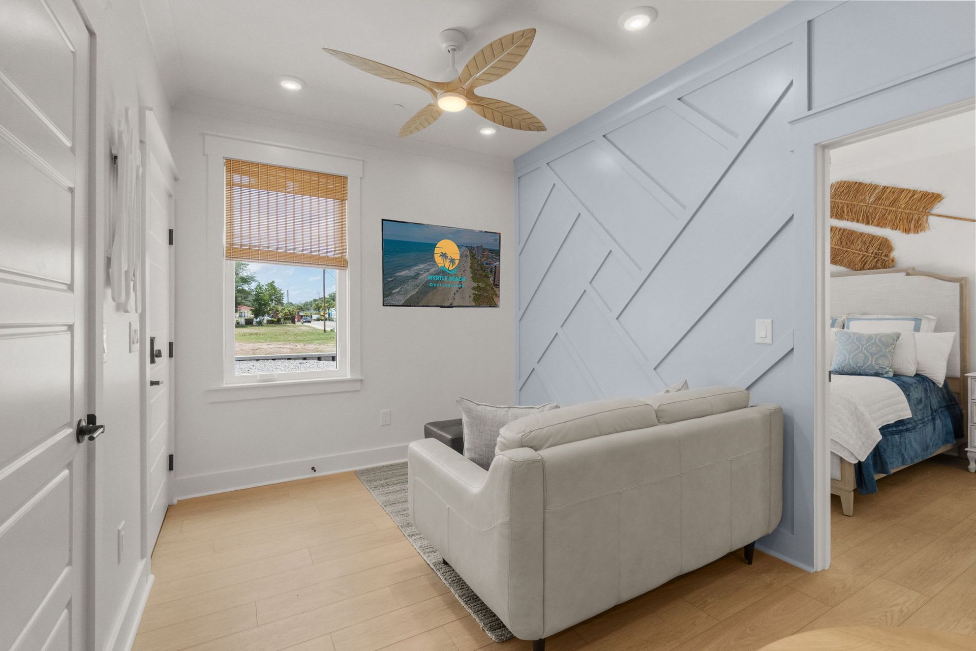 Bright bedroom with white walls, a sofa, window, ceiling fan, and blue accent wall leading to an adjoining room