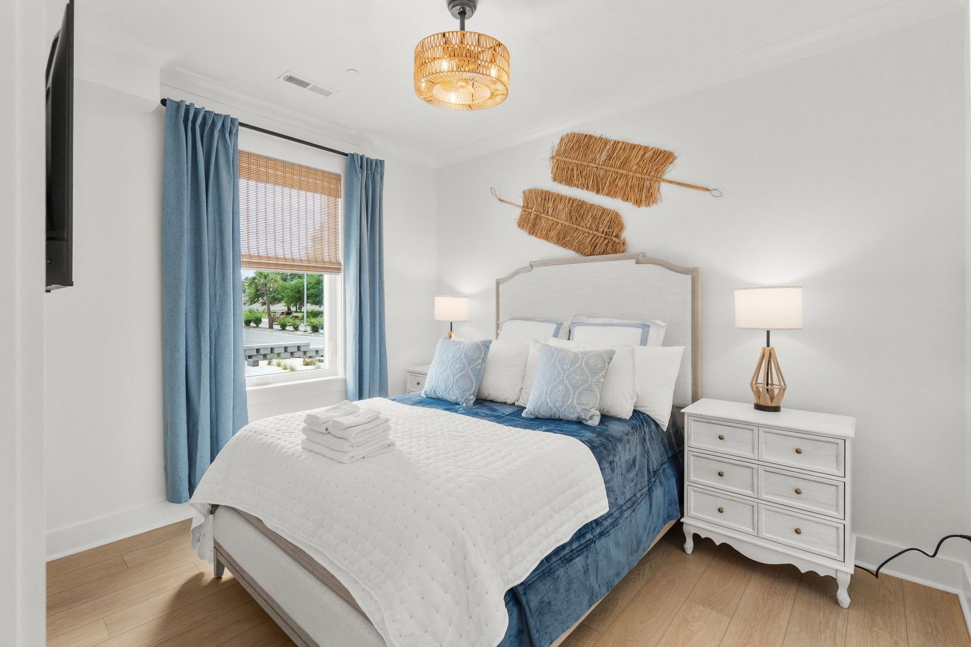 Bright bedroom with blue curtains, white bed, woven wall decor, and a patterned nightstand.