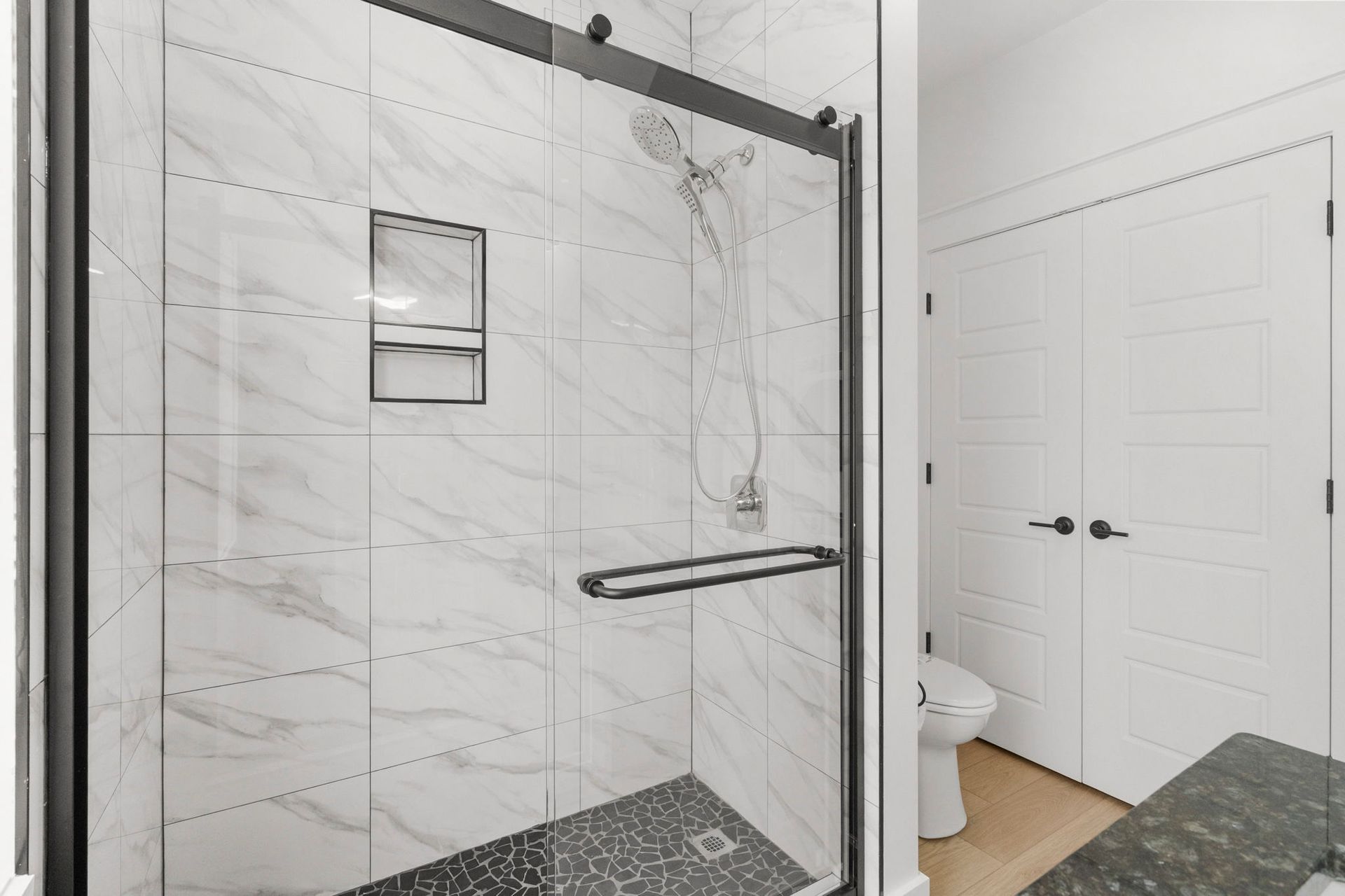 Walk-in shower with marble-look tile, black hardware, and a nearby bathroom door.