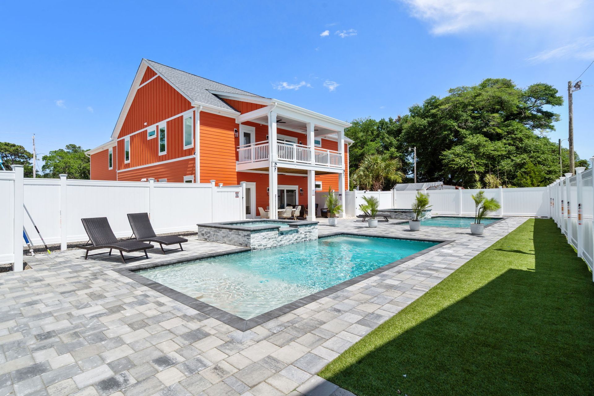 Backyard pool beside a red house with white trim and lounge chairs on a sunny day