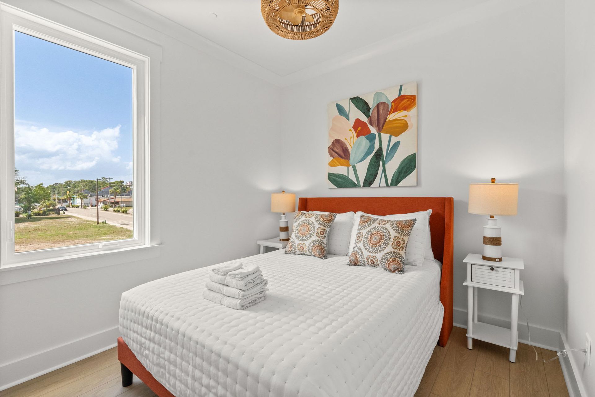 Bright bedroom with white walls, queen bed, bedside lamps, floral art, and a window view outside.