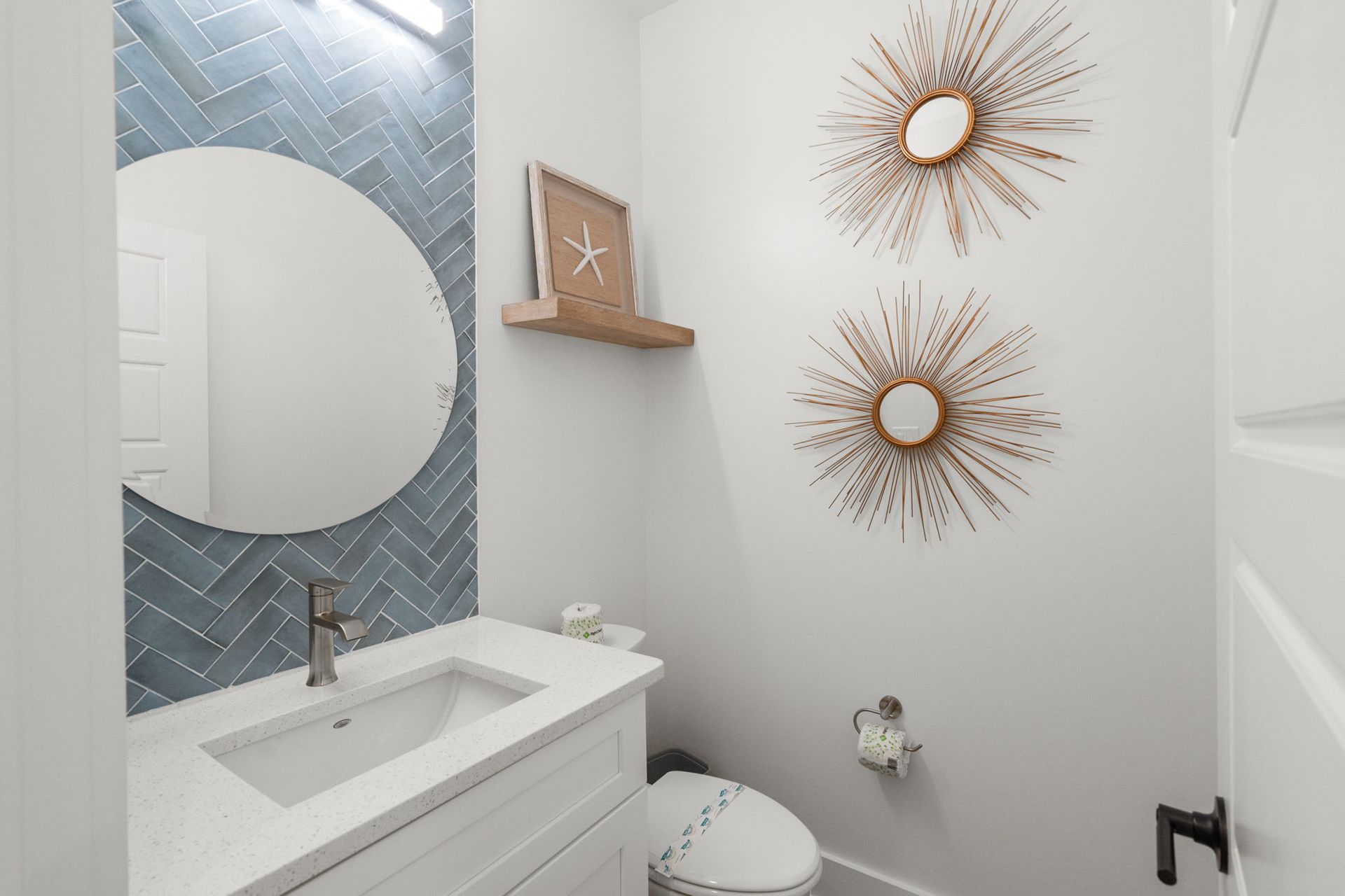 Small modern bathroom with white vanity, round mirror, blue tile accent wall, and gold sunburst decor