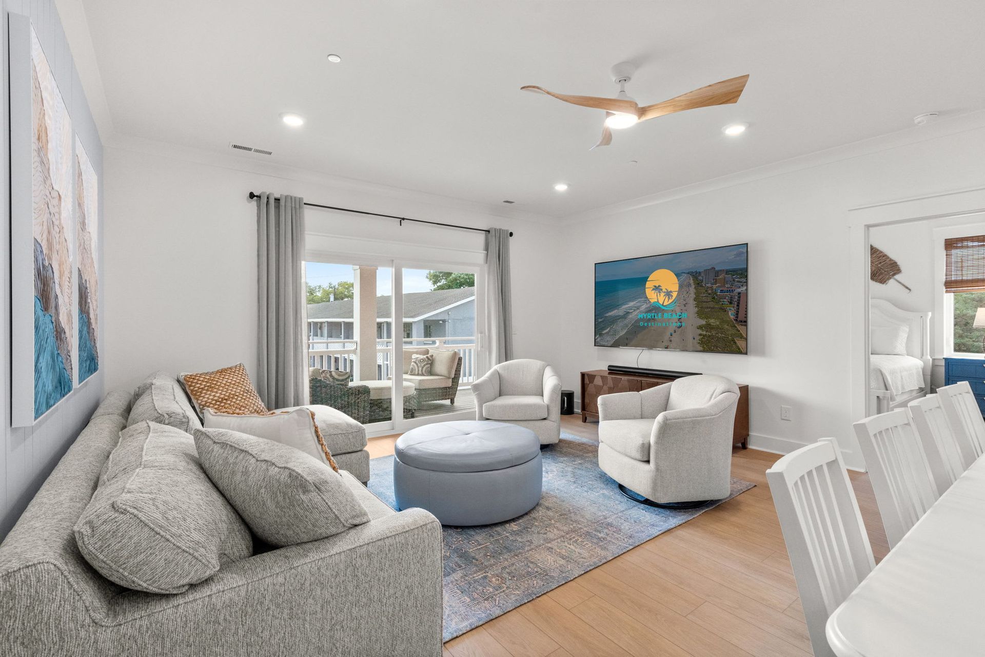 Bright living room with gray sofa, white chairs, blue rug, ceiling fan, and large windows to a balcony.