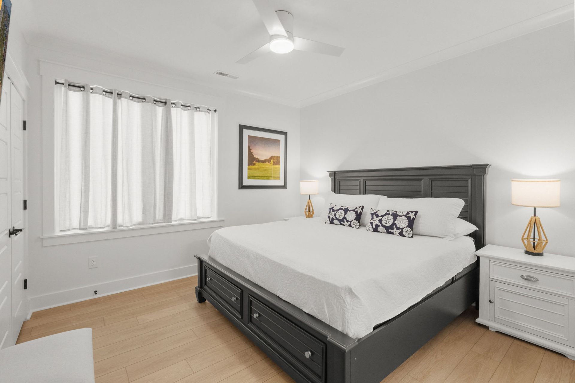 Bright white bedroom with a dark wood bed, nightstand lamp, window, and framed wall art.