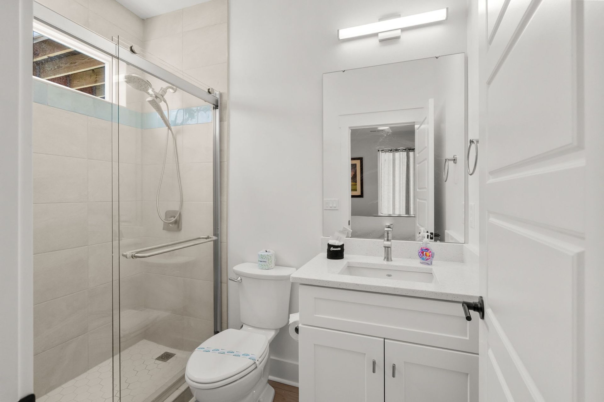Bright white bathroom with glass shower, toilet, vanity, and mirror under wall light.