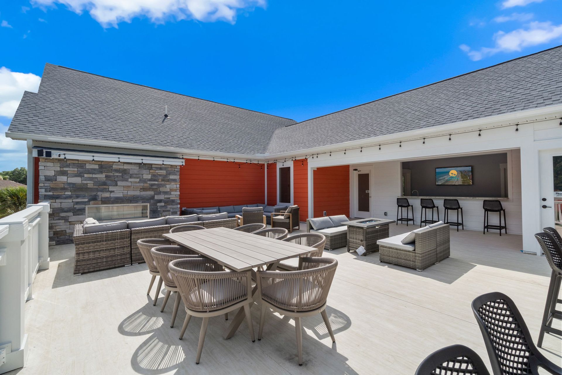 Spacious outdoor patio with wicker seating, dining table, stone fireplace, and an open kitchen under a blue sky