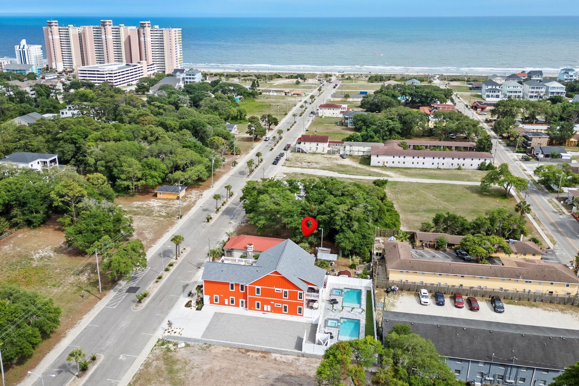 Aerial view of a coastal neighborhood with a red-roofed orange building, roads, trees, and ocean in the distance