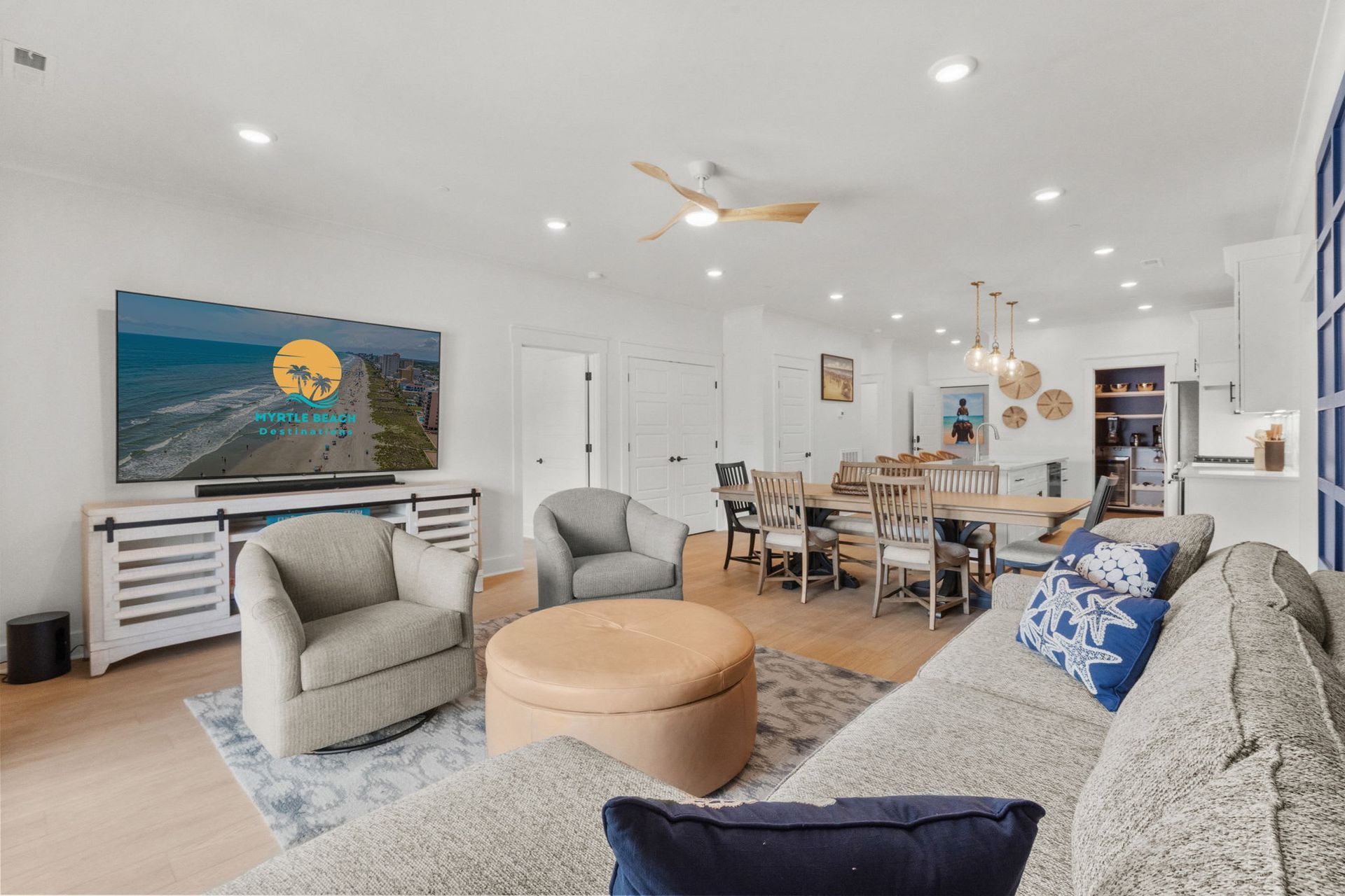 Bright modern open-plan living room and dining area with white walls, beige sofa, ottoman, and ocean artwork.