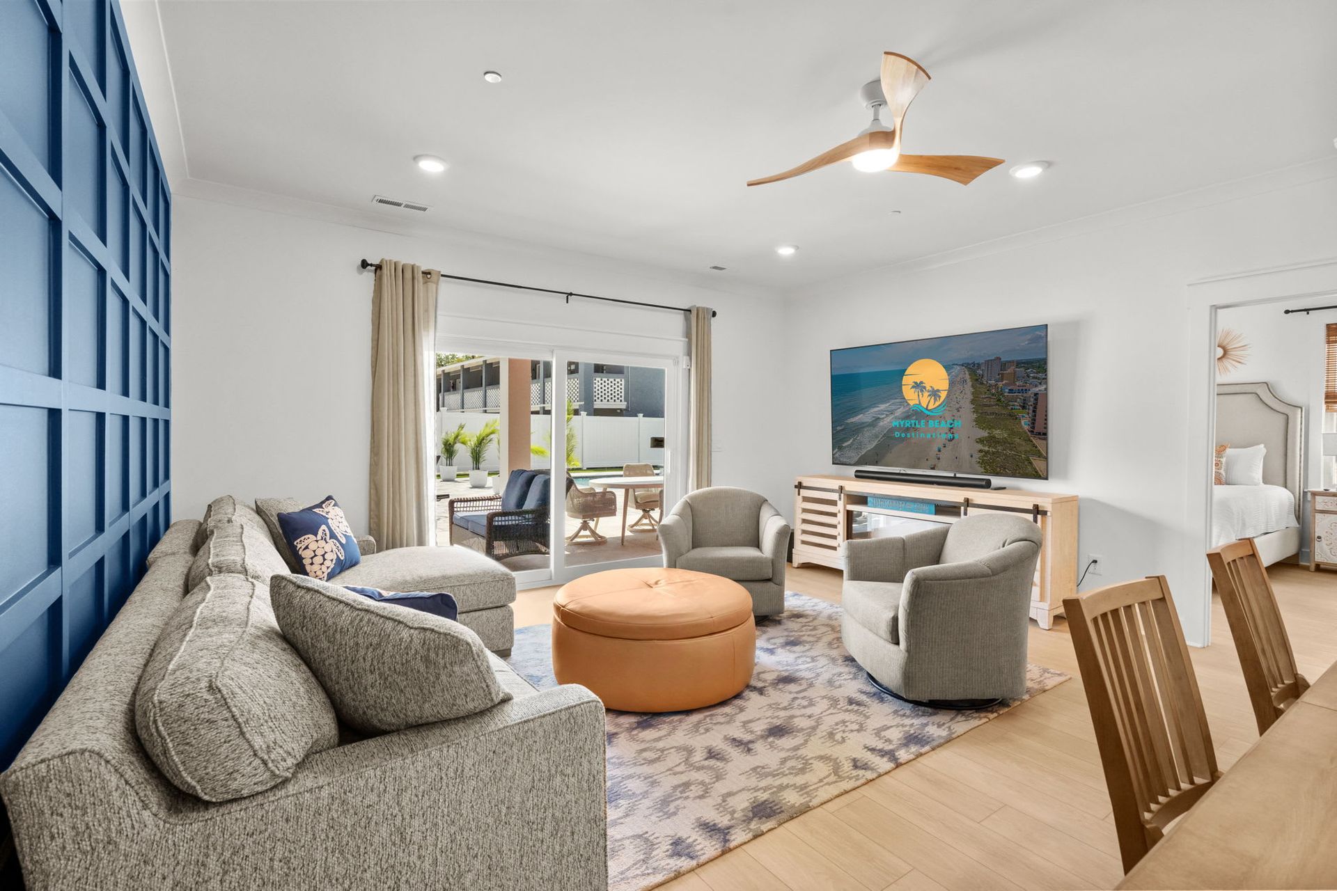 Bright living room with gray sofas, round tan ottoman, blue accent wall, and patio doors to a balcony