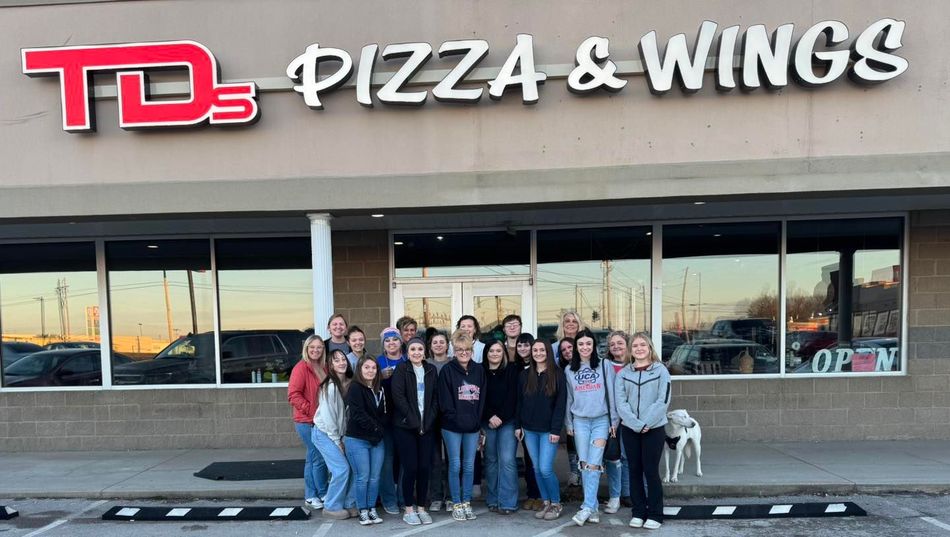 Group posing outside TD's Pizza & Wings storefront at dusk
