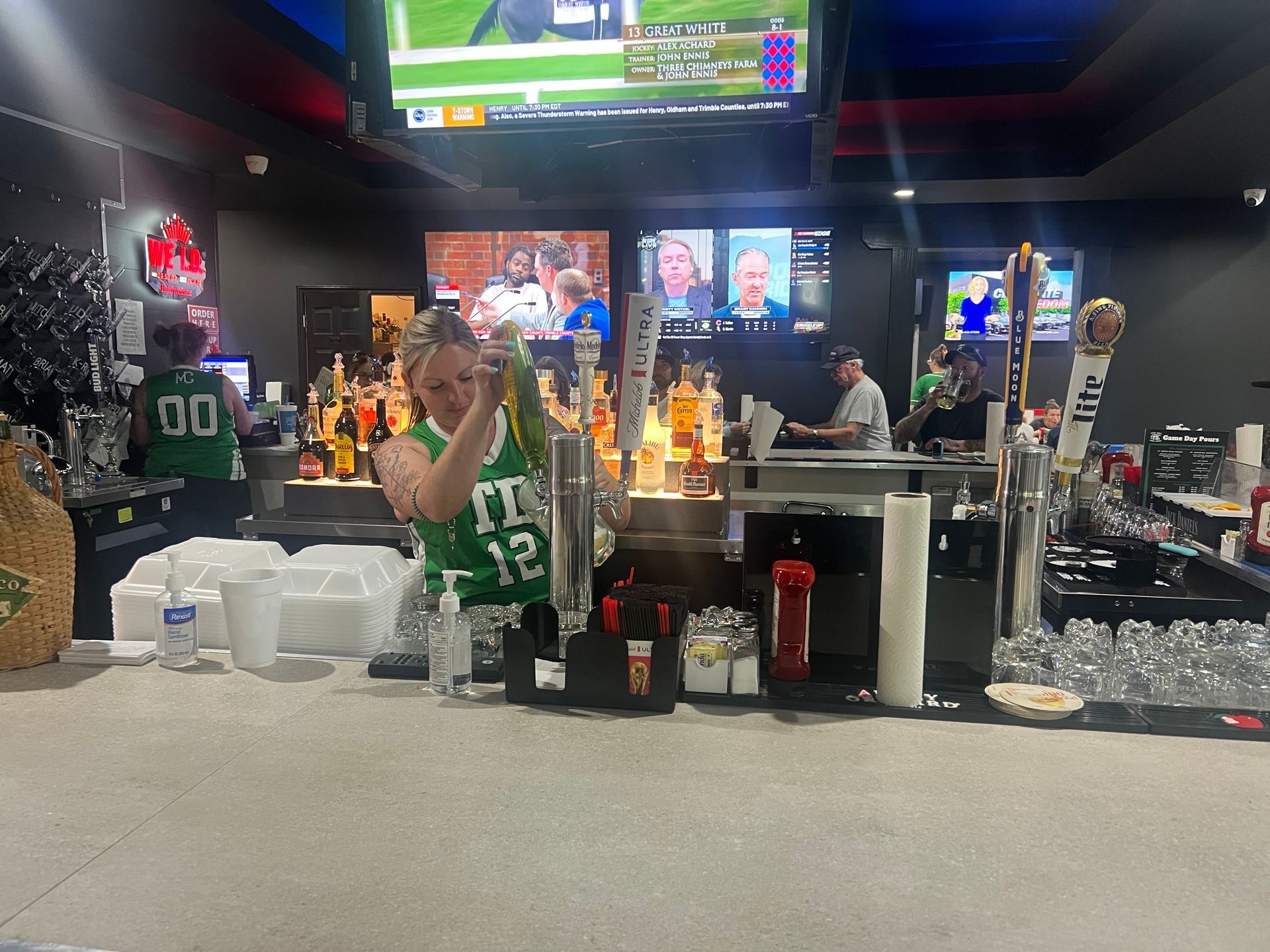Bar counter with drinks and TV screens; person in green shirt stands behind the bar