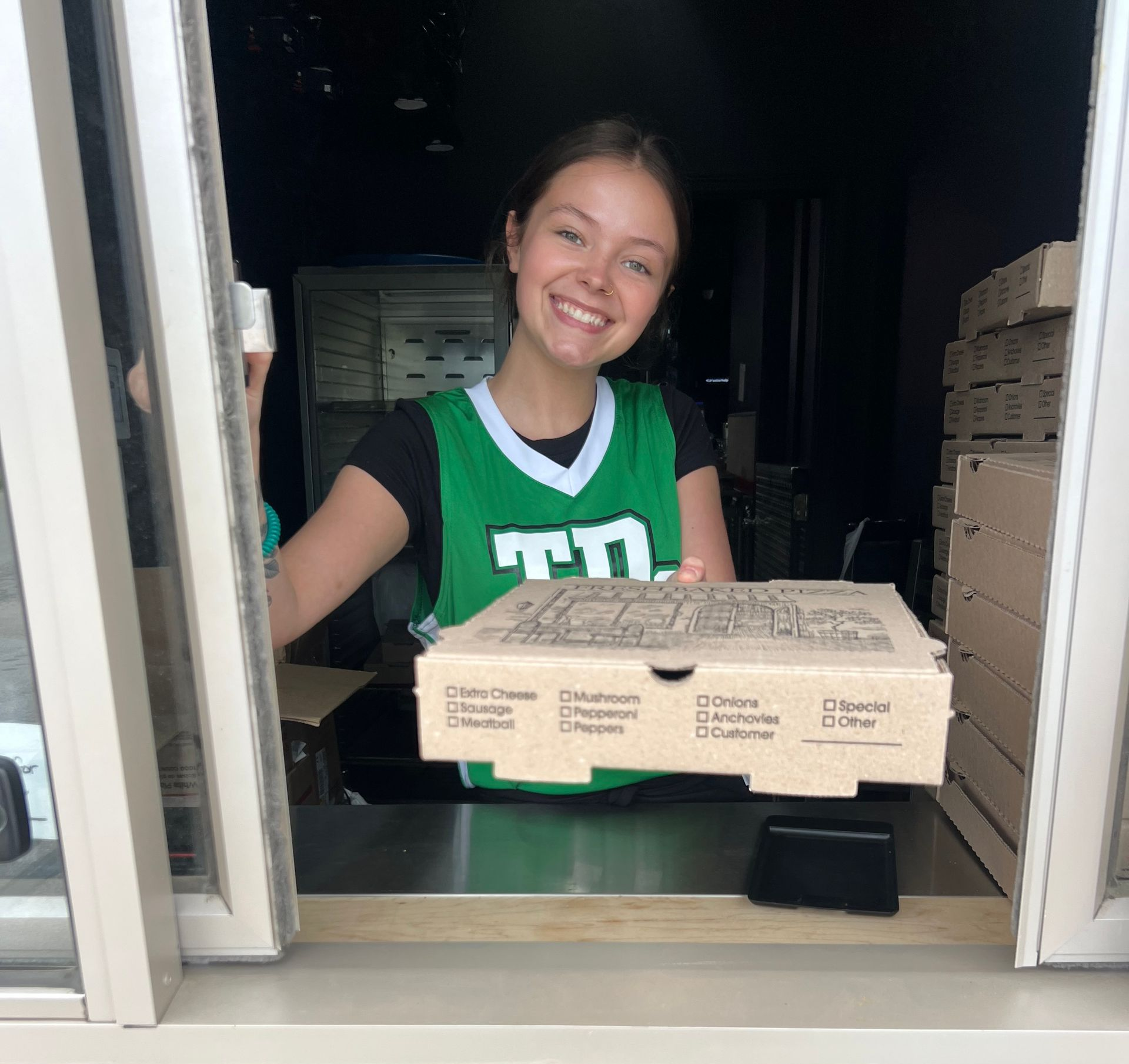 Smiling person in a green vest hands out pizza boxes through a service window.