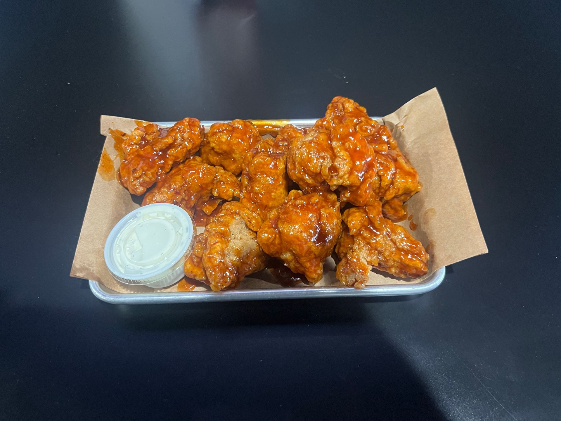 Tray of saucy buffalo chicken wings with a small cup of dipping sauce on parchment paper