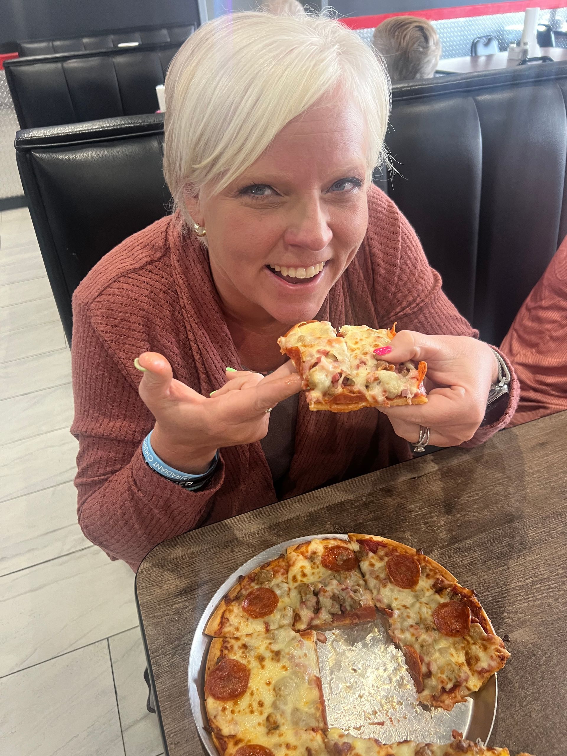 Woman in a restaurant holding a slice of pizza and smiling over a pizza on the table