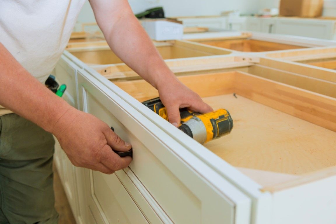 Hands installing a drawer with a yellow cordless drill in a light wood cabinet frame