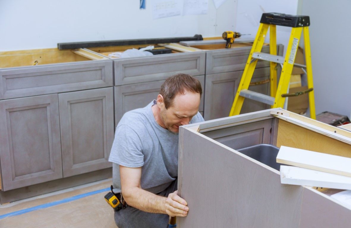 Man installing gray kitchen cabinets beside a yellow ladder in a partially renovated kitchen