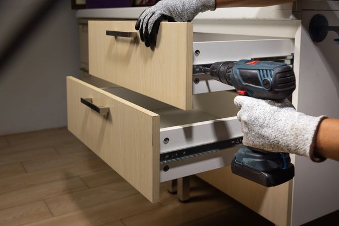 Gloved hands using a cordless drill to install a drawer in a light wood cabinet
