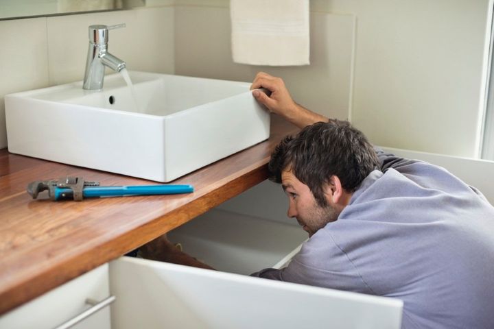 Plumber repairing a sink cabinet under a white bathroom basin, with blue tools on the counter.