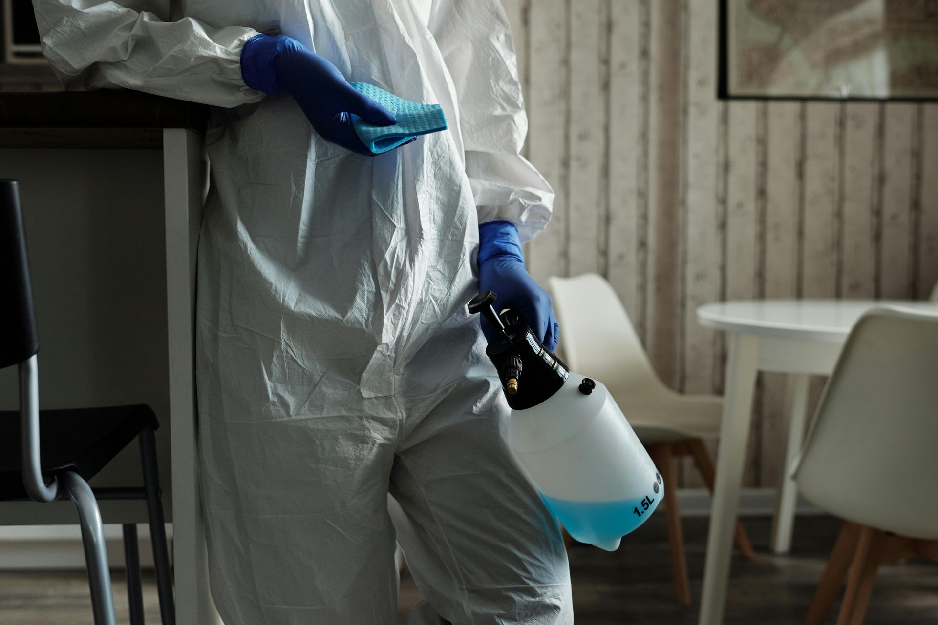Person wearing blue gloves holding spray bottles, with a yellow cloth and bucket on a surface.