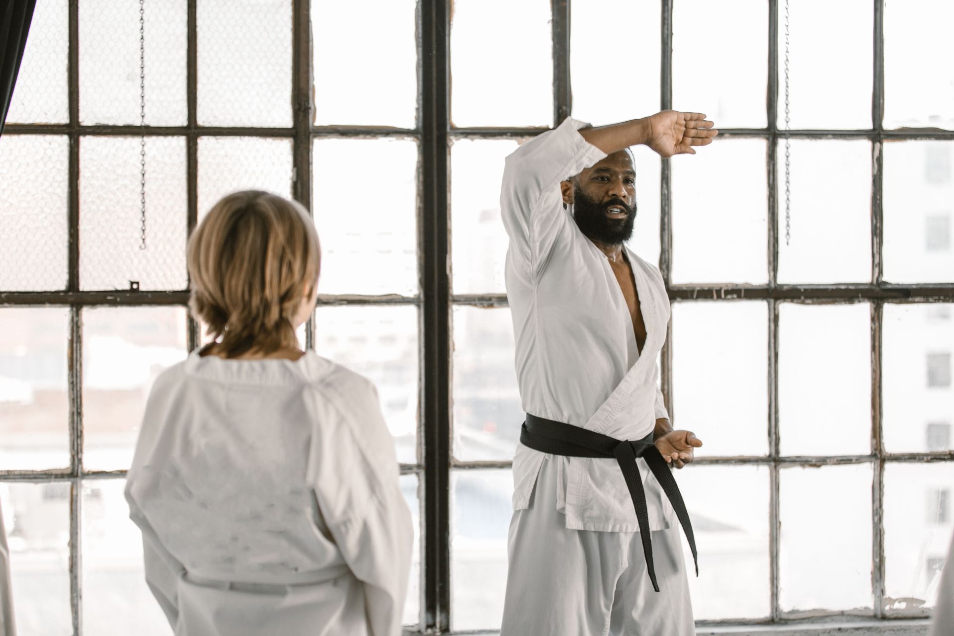 Two people in white martial arts uniforms practice in front of large windows.