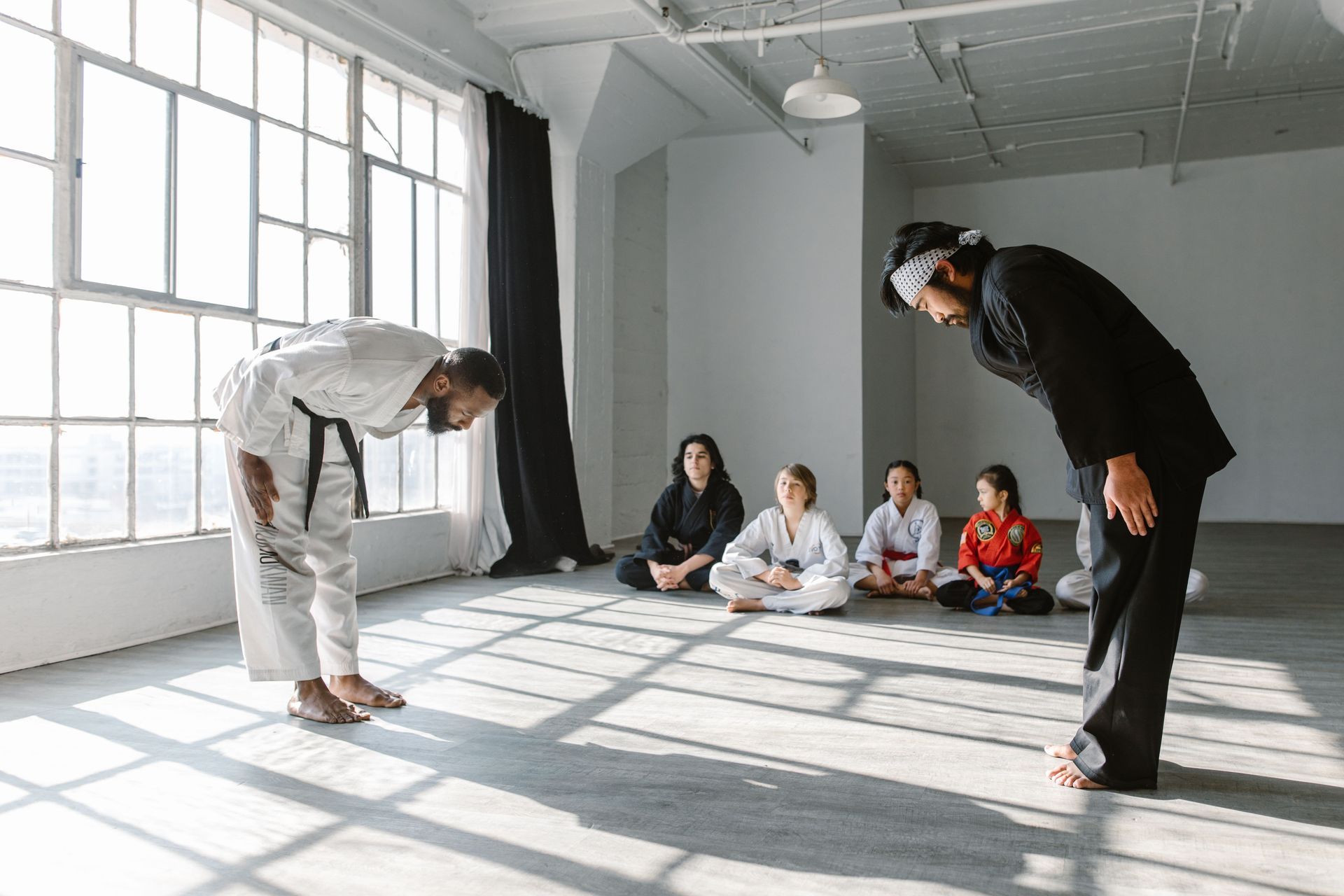 Two martial artists bow to seated students in a bright studio with large windows