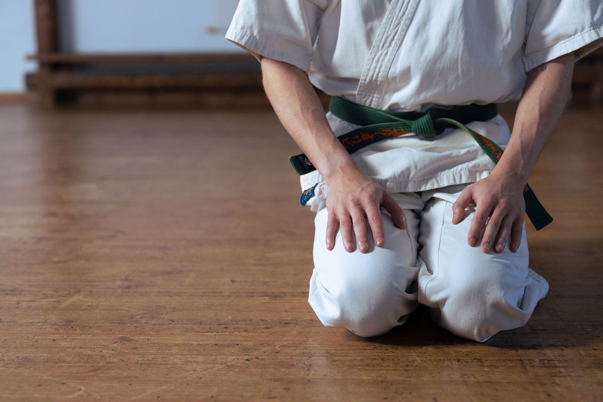 Person kneeling on a wooden floor in a white gi with a green belt, hands resting on thighs