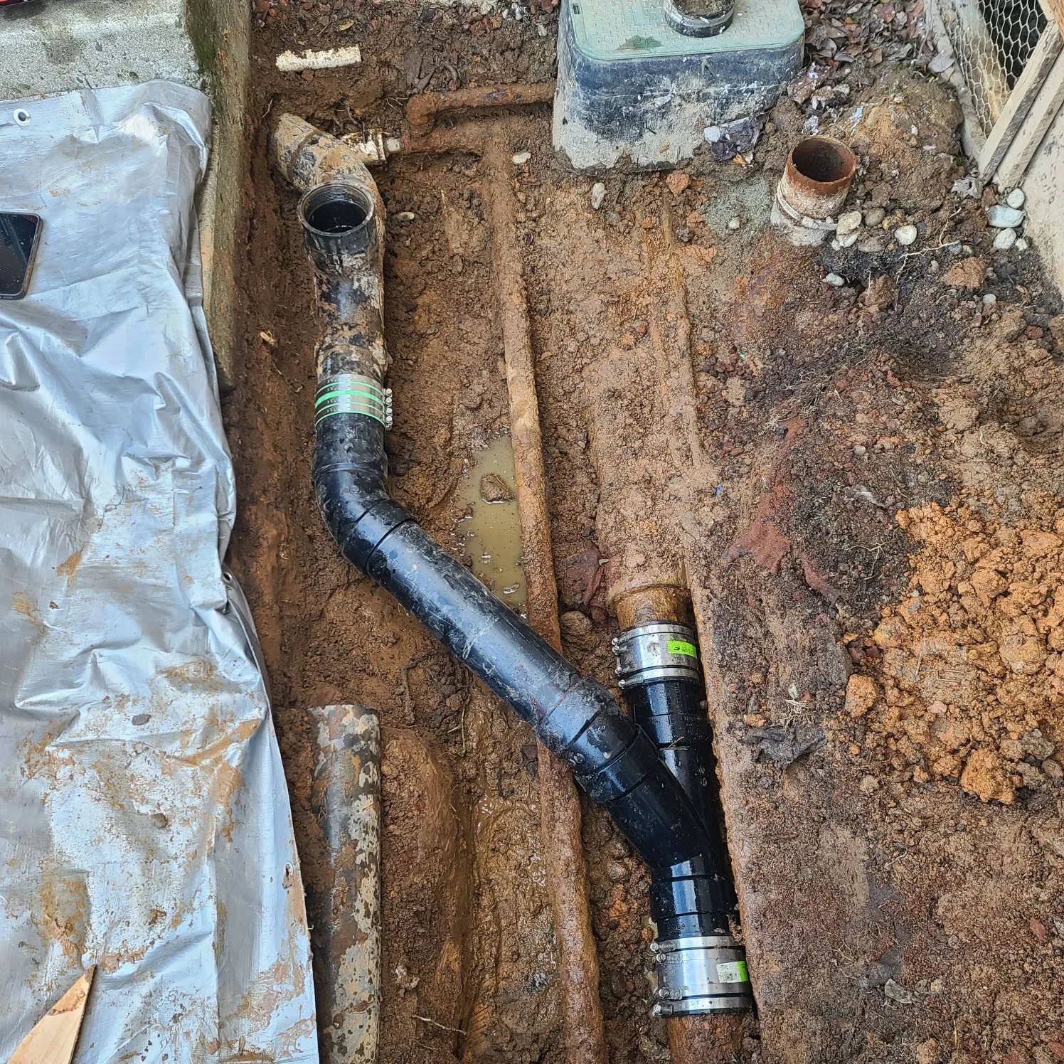 Black pipe connection in an excavated dirt trench beside a covered area.
