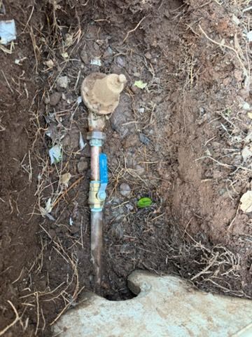 A brass water pressure regulator and blue-handled ball valve installed on a copper pipe in a dirt trench.
