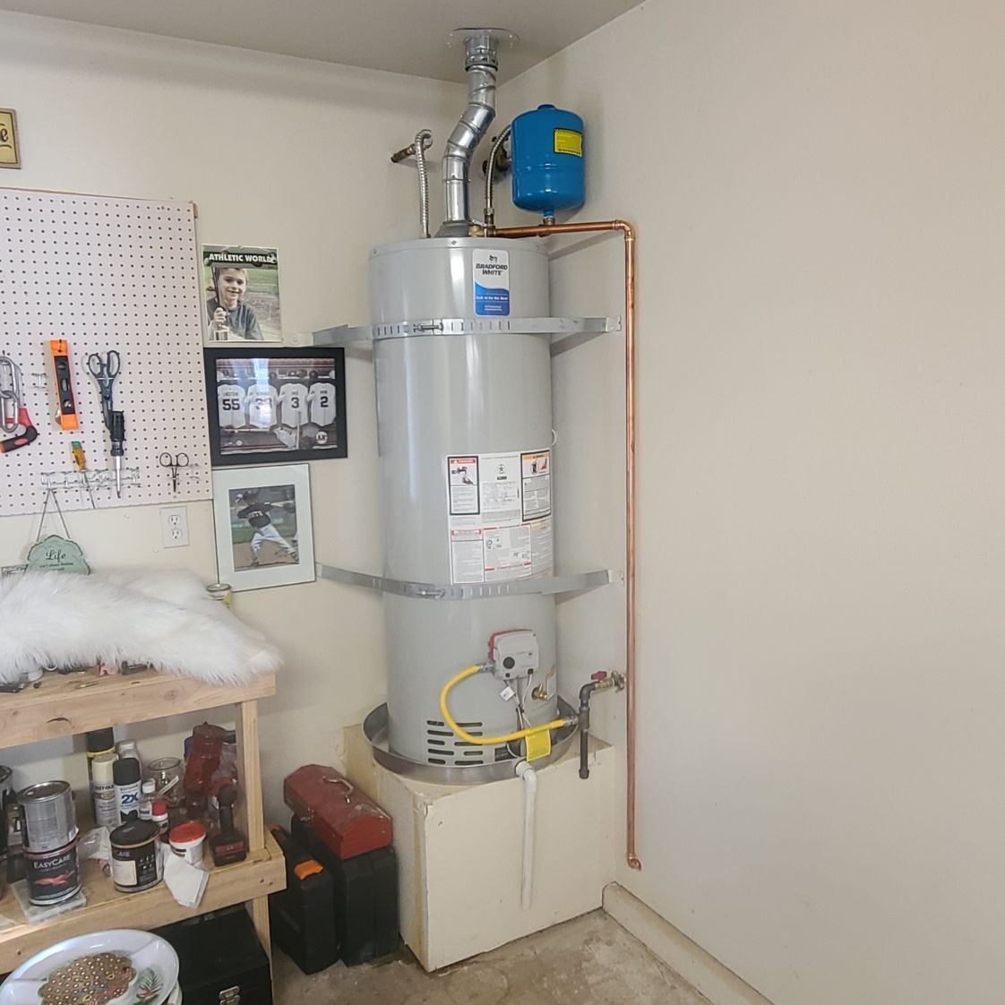 A water heater installed in a corner of a garage. It has a vent, copper pipes, and a blue expansion tank.