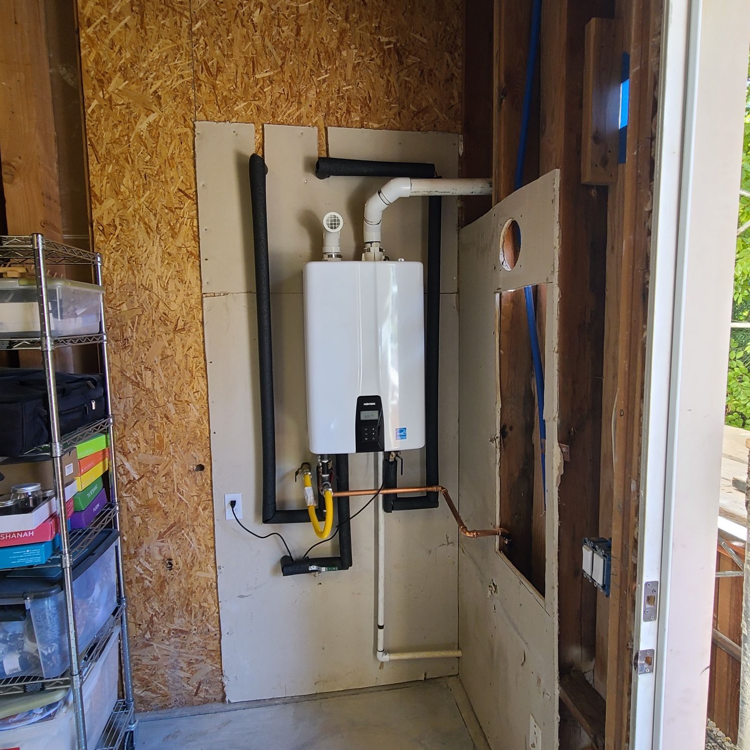 A wall-mounted, white tankless water heater in a utility room. Black pipes are insulated