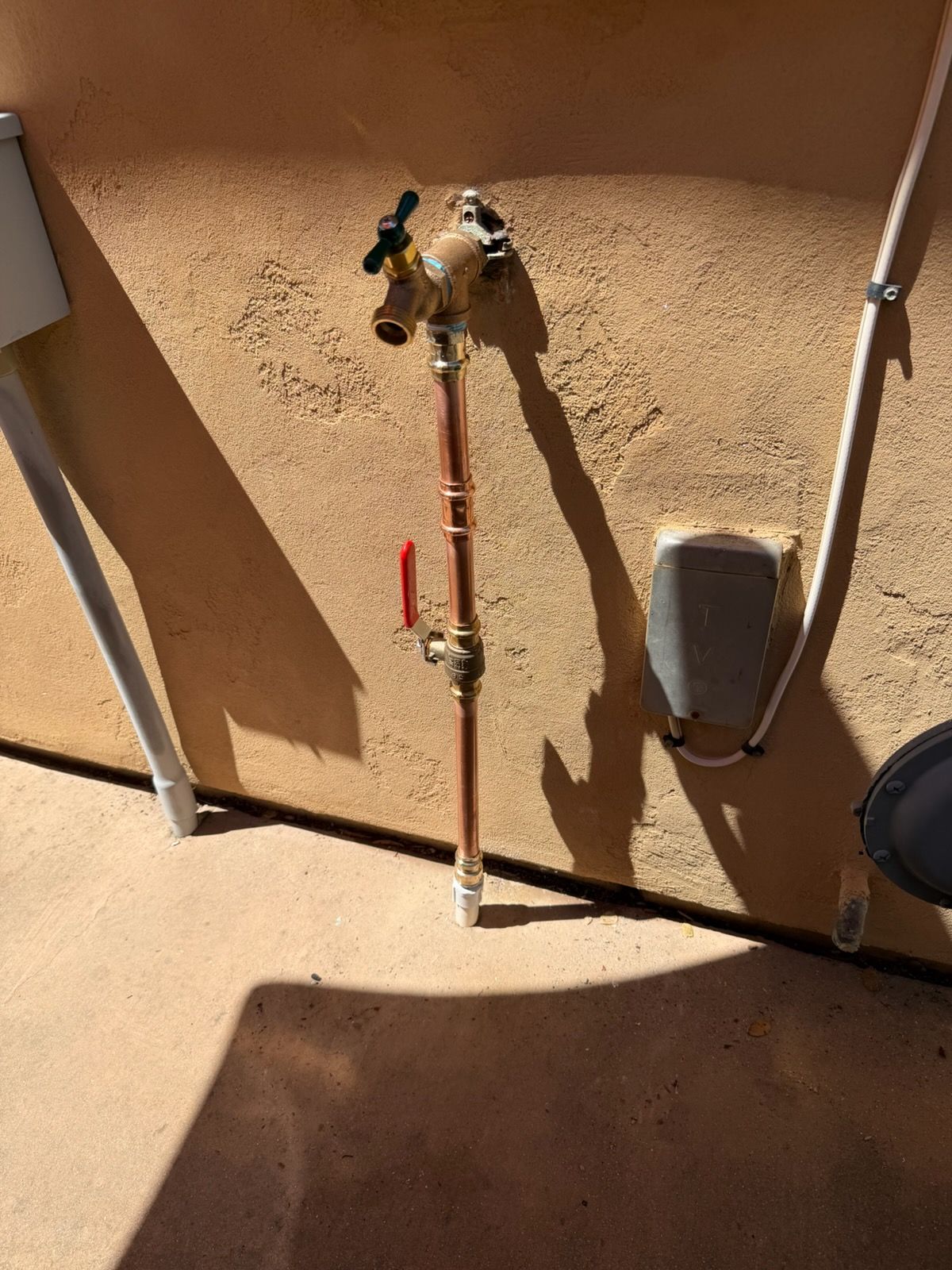 Outdoor wall with copper plumbing pipes, a red valve, and a small utility box in sunlight