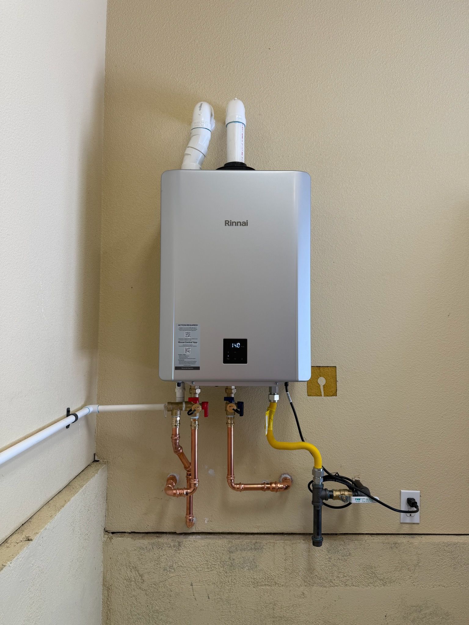 Wall-mounted white tankless water heater with exposed copper pipes and hoses in a utility room.