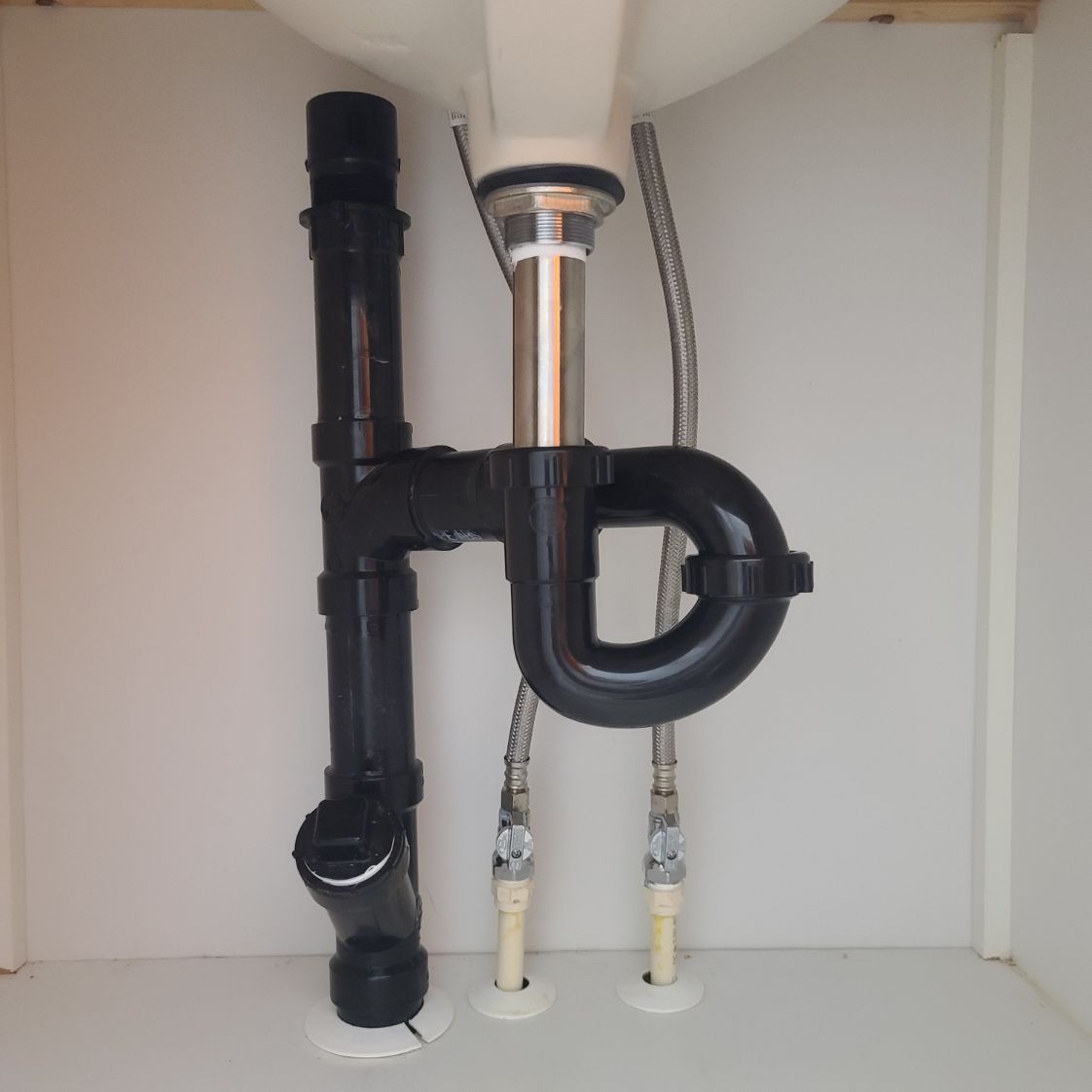 Black plumbing pipes under a white sink in a cabinet; includes a P-trap and supply lines.