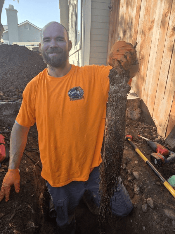 Man in orange shirt holding tree root, kneeling in a trench. Dirt, tools, and a wooden fence are visible.