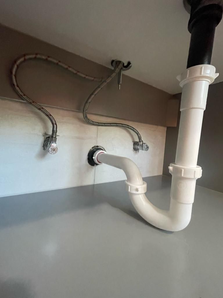 Under-sink plumbing: white pipes, silver braided hoses, gray countertop and cabinet interior.