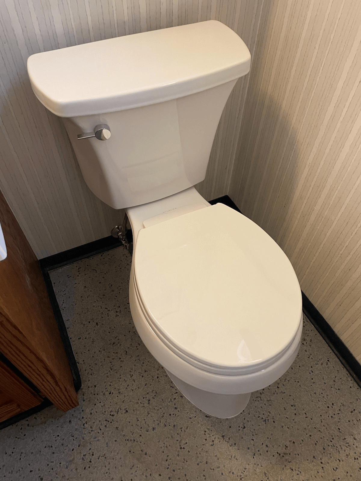 White toilet in a bathroom, with a closed lid. The floor is speckled, and the wall has wallpaper.