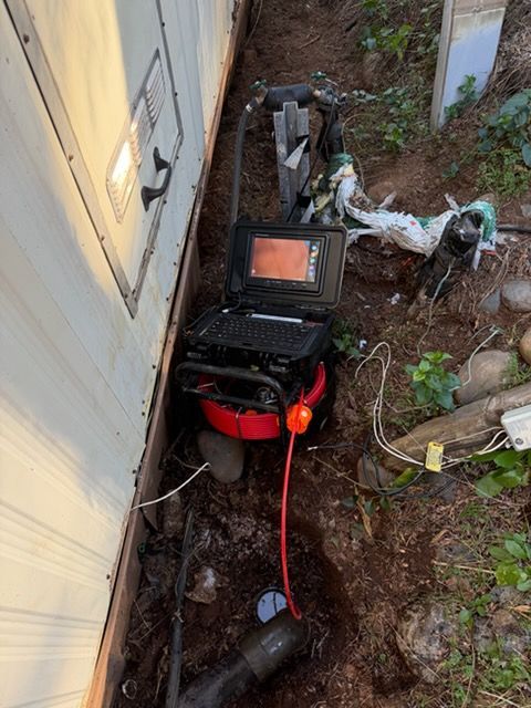 A sewer inspection camera system with a monitor and red drum is inserted into a pipe in the ground next to a mobile home.
